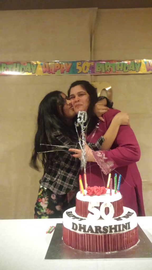 A lady with long dark hair and black check shirt embraces another lady wearing a maroon dress looking at the camera. There is a “Happy 50th Birthday” banner in rainbow colours behind them on a cream wall and a large two-tiered maroon and white birthday cake with “50 Happy B’day Dharshini” written on it