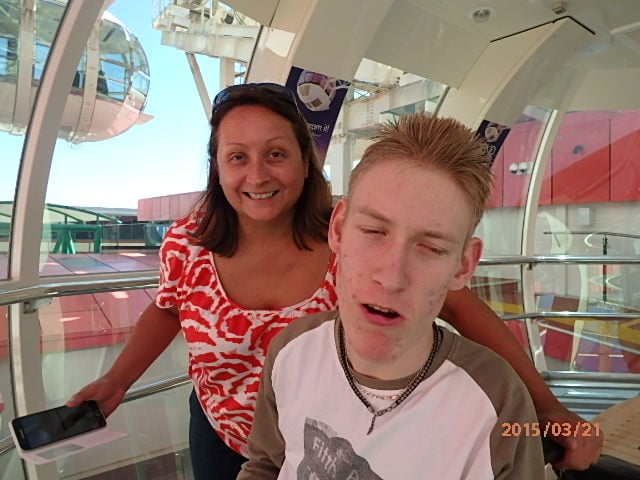 Two people pose for a photo within a Ferris wheel pod. The young man to the front of the picture smiles with his eyes partially closed, and a woman in a bright red and white top stands behind him. Another Ferris wheel pod can be seen through the window to the side of the pair.