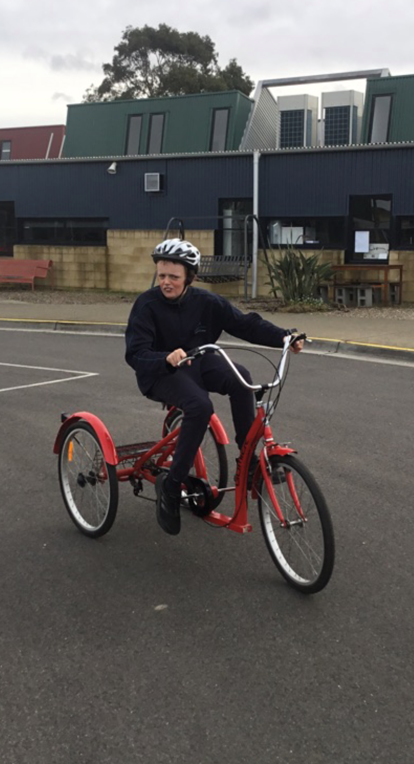 A person in a dark tracksuit and white helmet sits on a red tricycle looking to their right. They are on a road with a dark blue and green building behind them