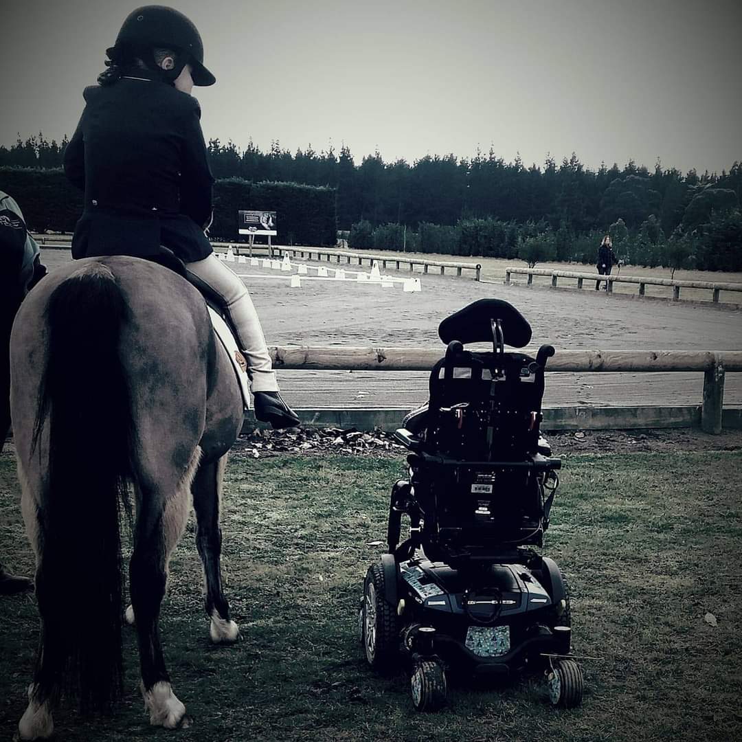 back view of a young person in an equestrian rider’s jacket and peaked helmet sits astride a grey horse. The horse rider is looking to their right at their power wheelchair. The rider and horse are looking out towards an equestrian training area