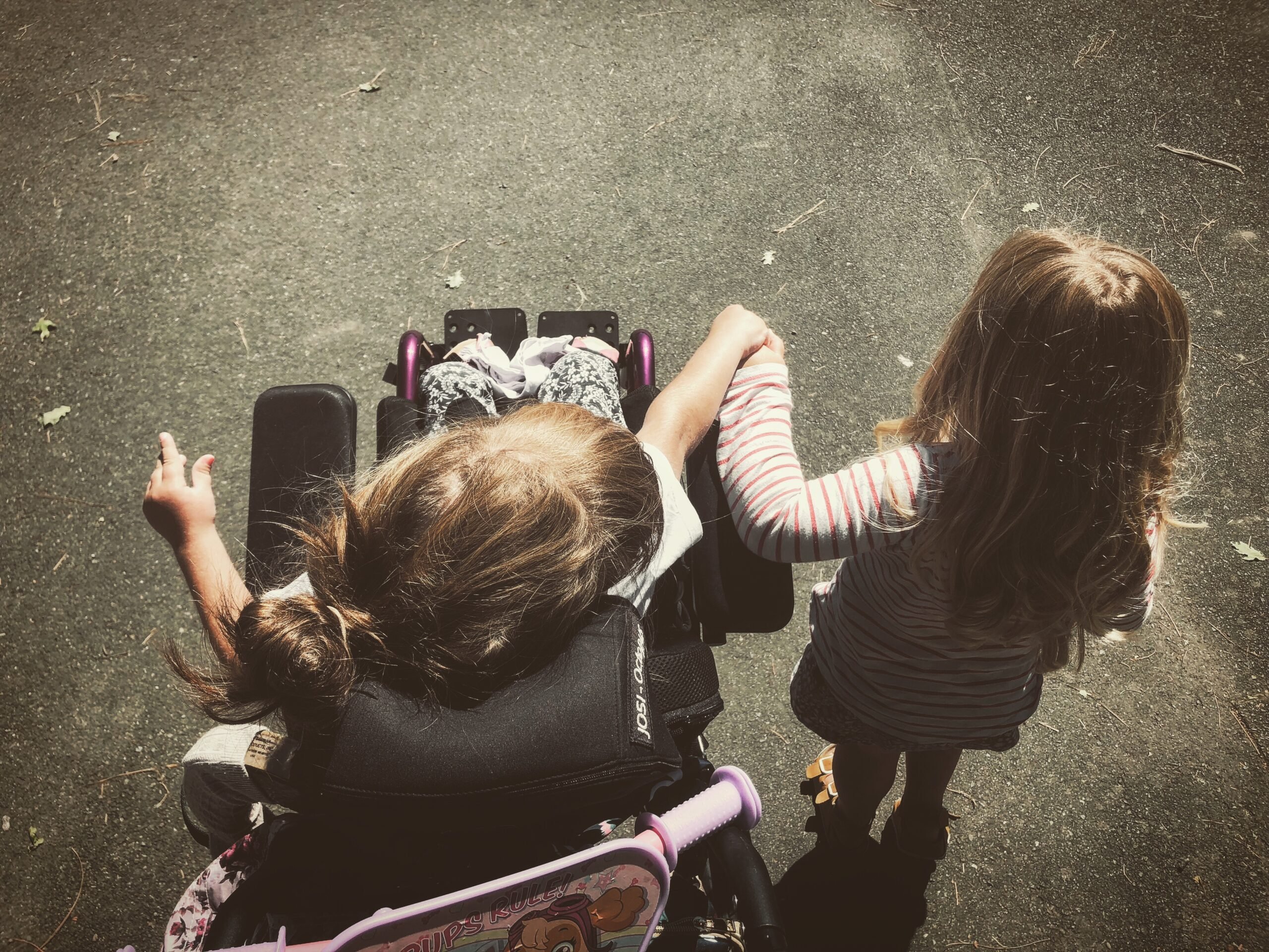 A bird’s eye top-down perspective shot that shows a child on the right in a wheelchair with her head turned towards, and right hand reaching out and holding onto a girl’s hand walking beside her. They are walking on black concrete ground