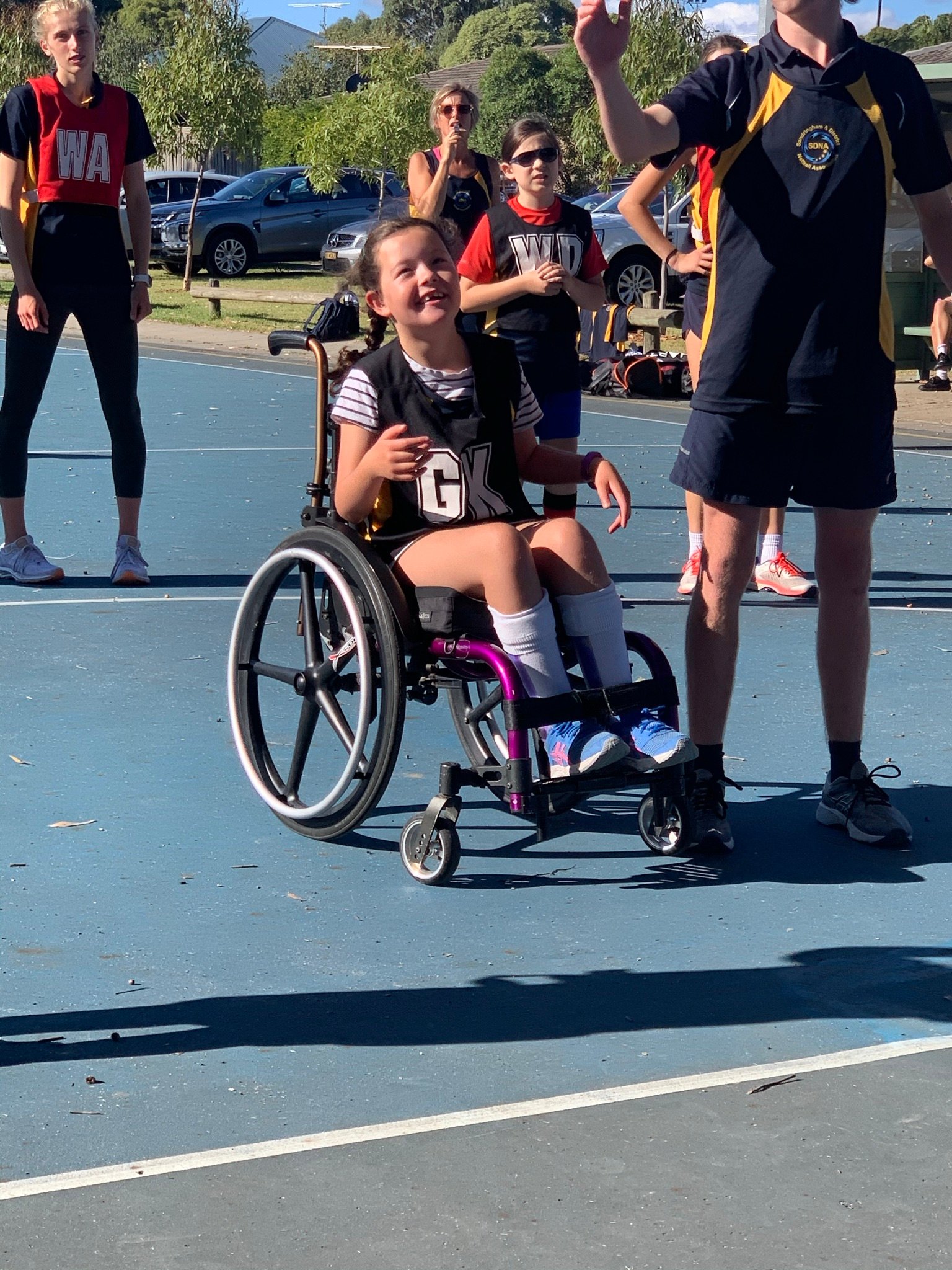 A girl in a “GK” netball bib sits in a purple wheelchair on a blue netball court smiling and looking up. There is a person in a navy t-shirt standing next to her pointing up, people in a “WA” and “WD” bib stand behind a white line behind her in the background