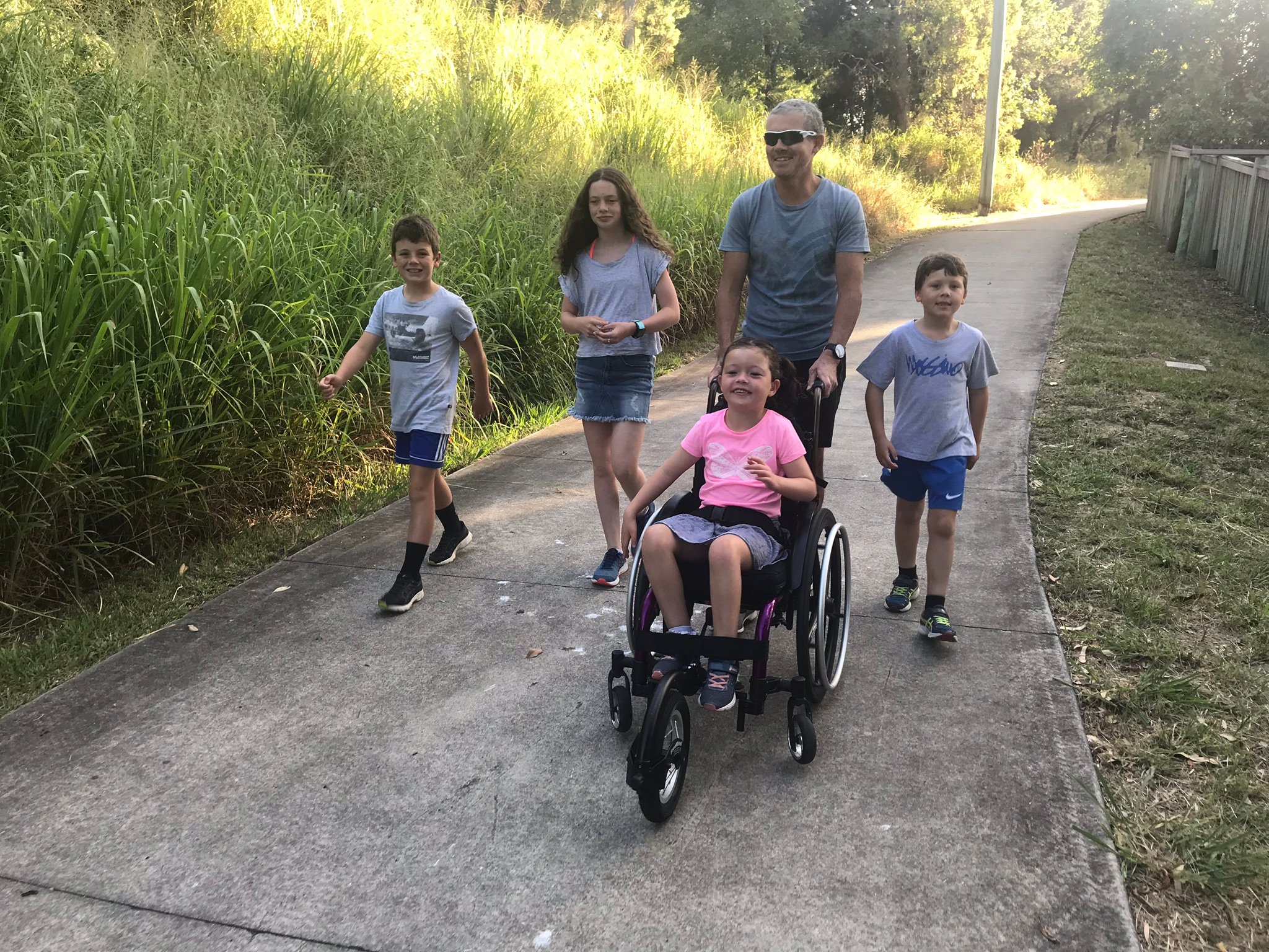 A girl in a pink t-shirt and purple wheelchair with an added front wheel smiles while being pushed by a man in sunglasses and a grey t-shirt. A young boy and girl, also in grey tops walk on the man’s left and another rboy in a grey top walks on the man’s right. They walk on a concrete path with a slope of overgrown grass to one side and flat patchy grass to the other side