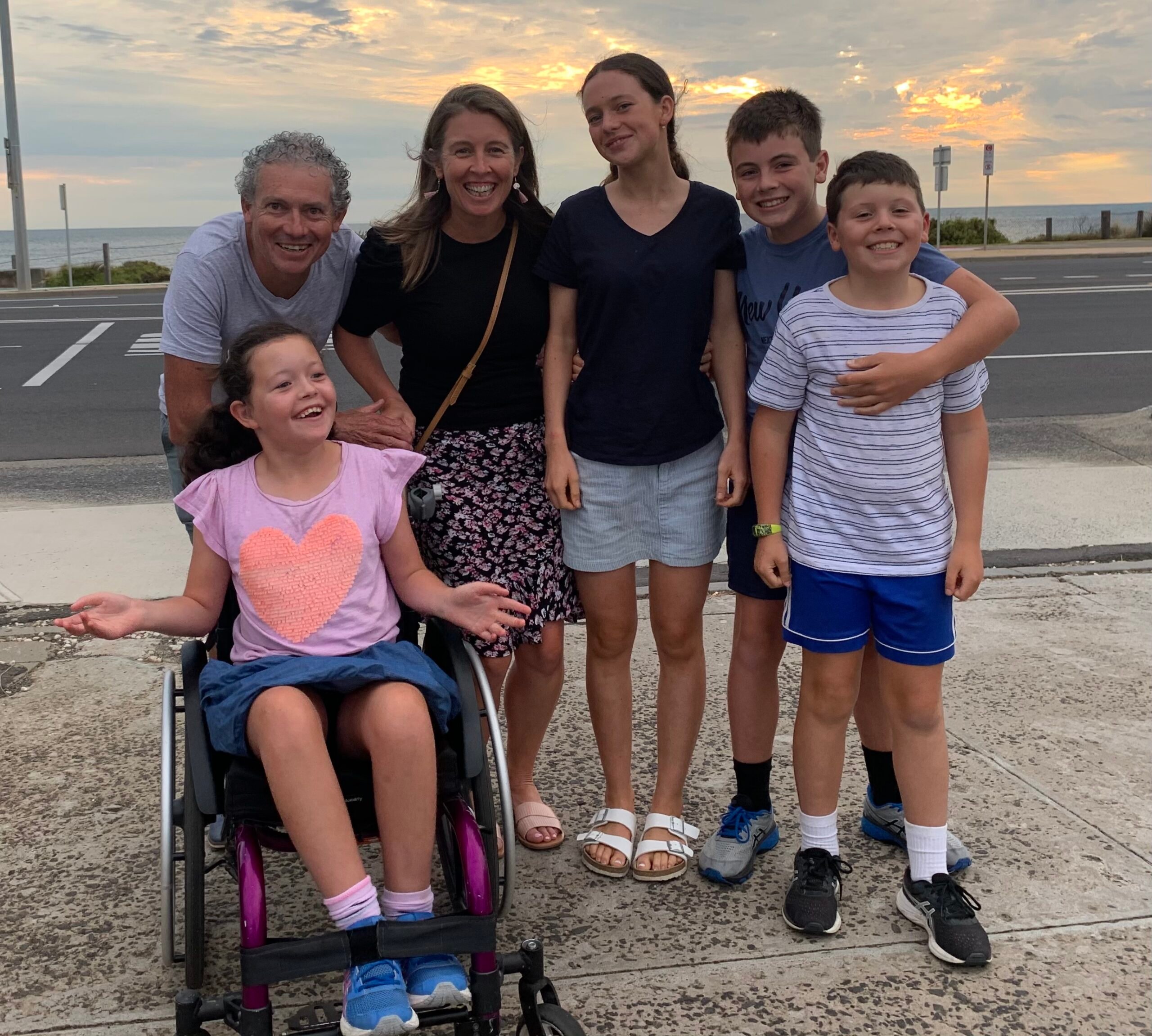 A family of six pose together for a photo: a girl in a pink top in a purple wheelchair, a man leans on her wheelchair behind her, holding the hand of a woman in a black top and three other children standing to the side of them. A road and beach in the background with a sun setting behind a cloudy sky.