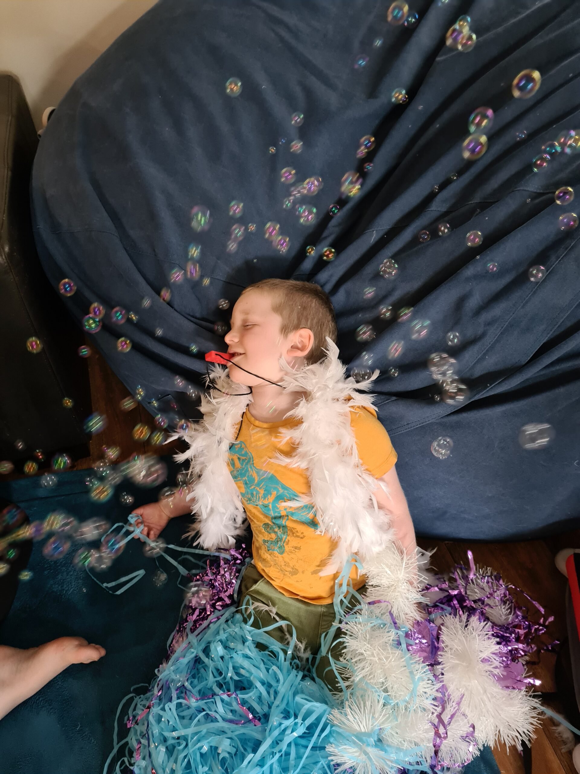 a young boy in a yellow t-shirt draped in a white feather boa scarf and blue, white and purple sparkley tinsel across his lap lies with his head on a dark blue bean bag and bubbles floating across his body