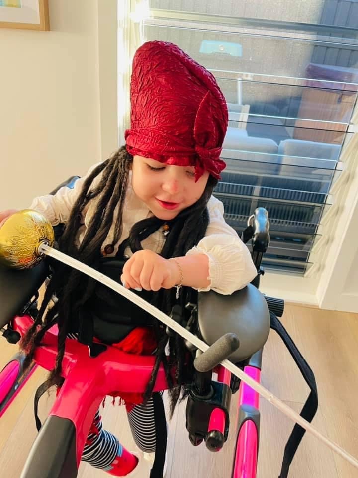 a young child in a fancy red pirate bandana hat with black dread locks spilling out around behind the hat stands in a pink walker. The child holds a fake sword and is looking down at the ground pointing the sword downwards