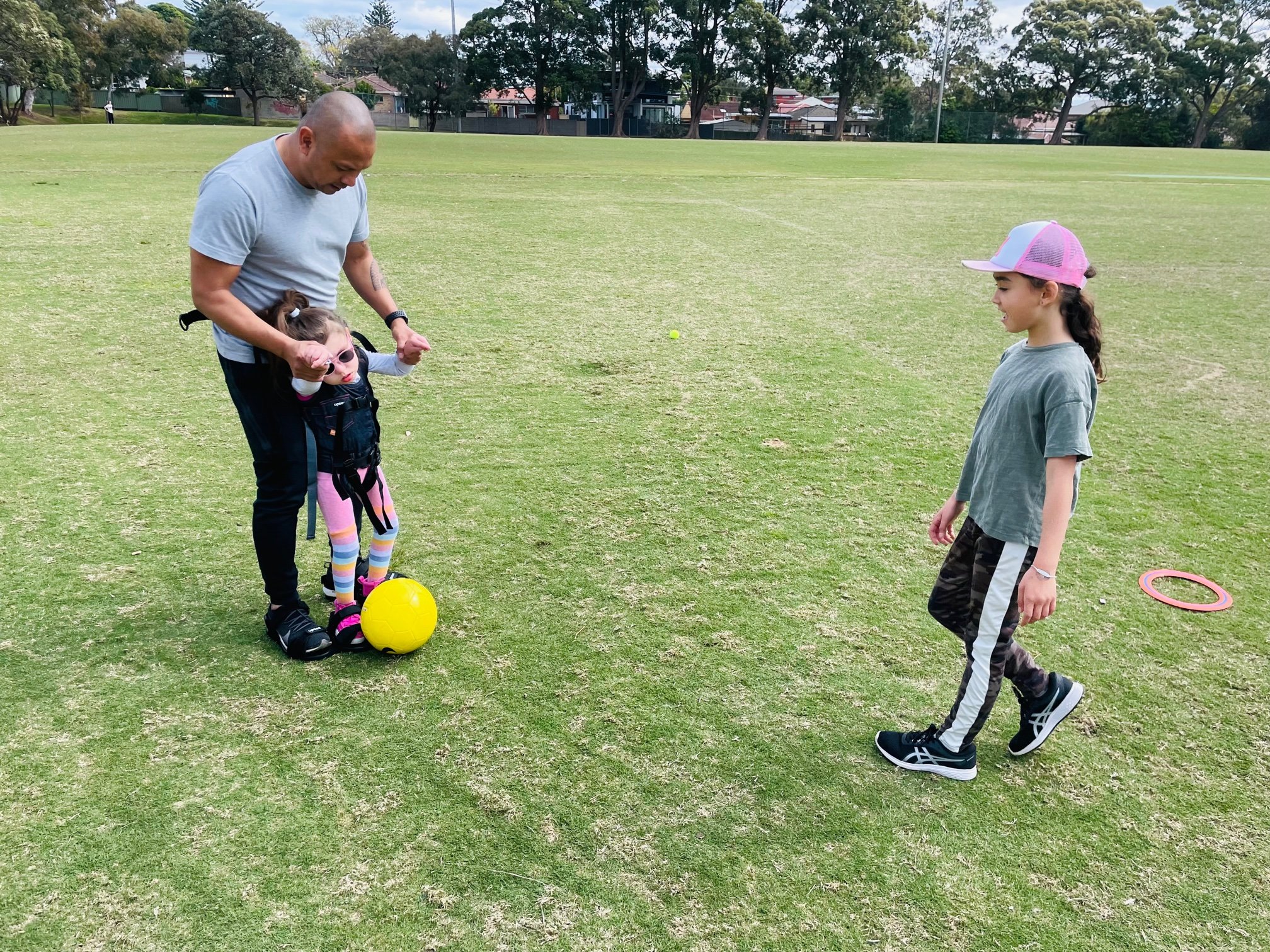 A young girl in pink leggings, rainbow socks and a vest with lots of straps stands while her hands are held by a man in a grey shirt behind her. There is a bright yellow ball at her feet. Another taller girl stands opposite her as if walking towards her. They are on a vast grassy field with trees and houses dotted in the background.