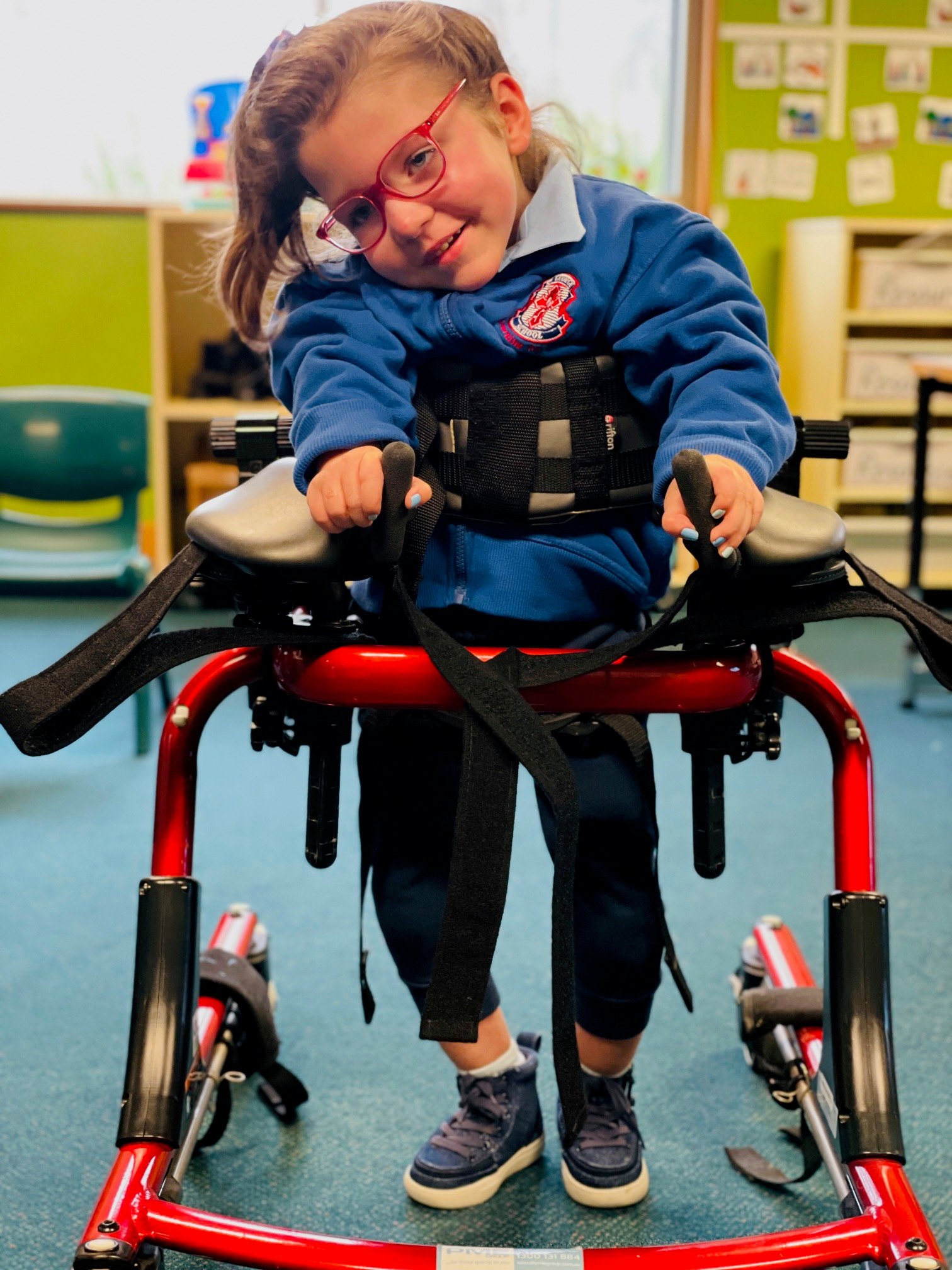 A young girl in a blue school uniform and red glasses stands and smiles in a red walking frame within a classroom with green walls