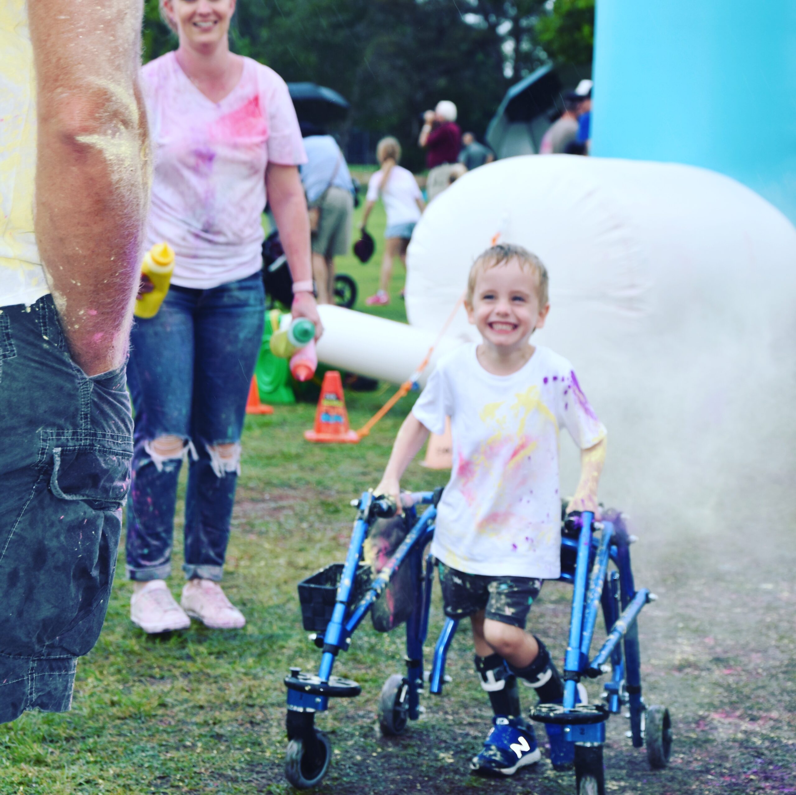 a young child walks with a blue metal walker with a wide smile on their face. They are wearing a white t-shirt covered in bright paint with a large inflatable ball behind them. An adult (also wearing a white t-shirt covered in paint) stands just behind the child. A person’s hand in their pocket (arm covered in paint) borders the left of the picture