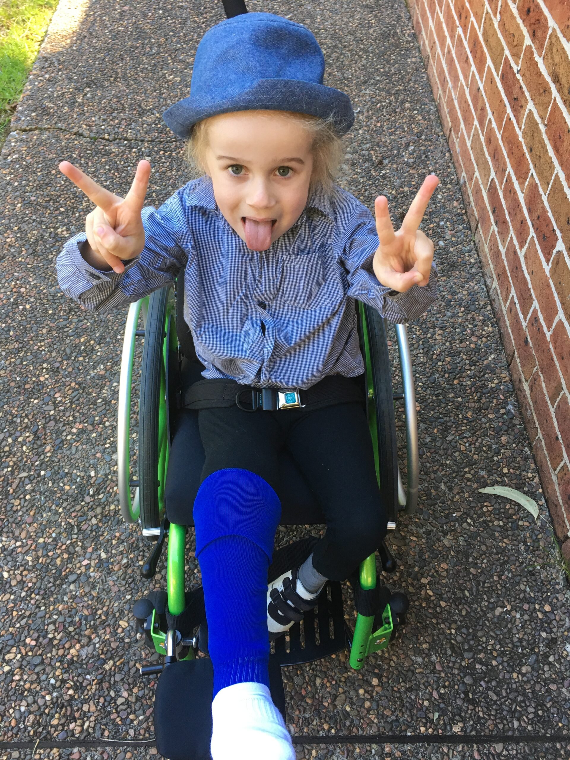 a young child in a blue shirt and hat and matching blue sock sits in a green wheelchair on a concrete path with the blue socked foot elevated. They poke their tongue out and hold two “peace” signs with their hands