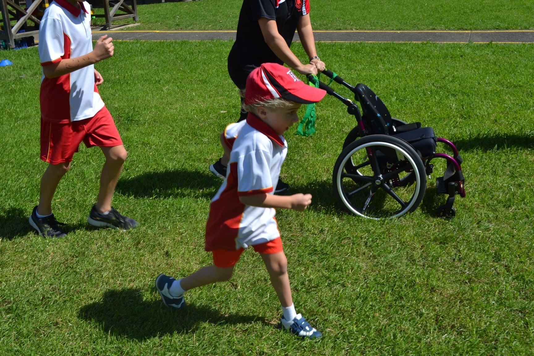 A young child in a red and white sports uniform and cap runs on a grassy field. An adult runs beside him pushing and empty wheelchair and another child runs behind them, the other people’s heads are cropped out of view