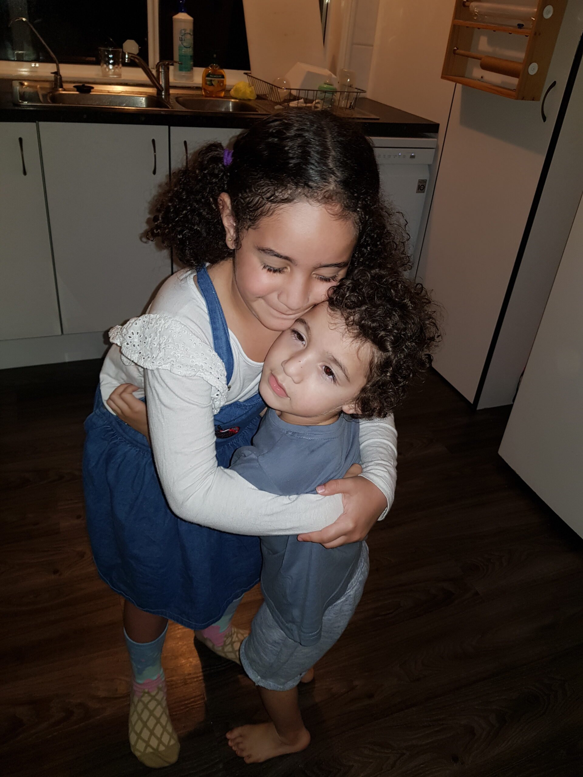 A girl crouches over with her eyes closed to cuddle a younger boy who leans up to cuddle her back. They stand in a kitchen setting