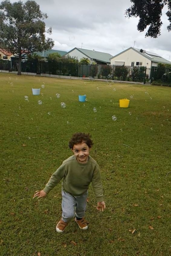 a young boy in an olive green jumper walks and smiles towards the camera with a field of grass and three coloured buckets behind him. Bubbles float above the boyd in the background. Houses can be seen beyond the field of grass