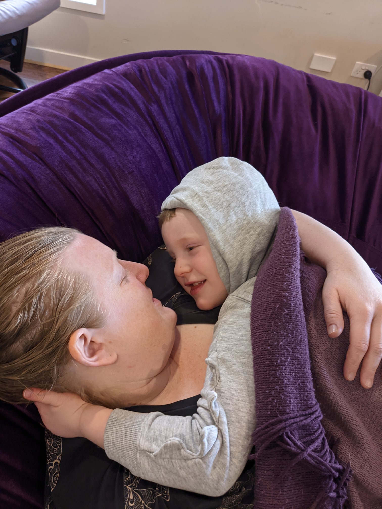 An adult sitting on a purple settee looks towards a child in a grey hoodie jumper wrapped in a purple blanket. They are looking at each other’s faces.