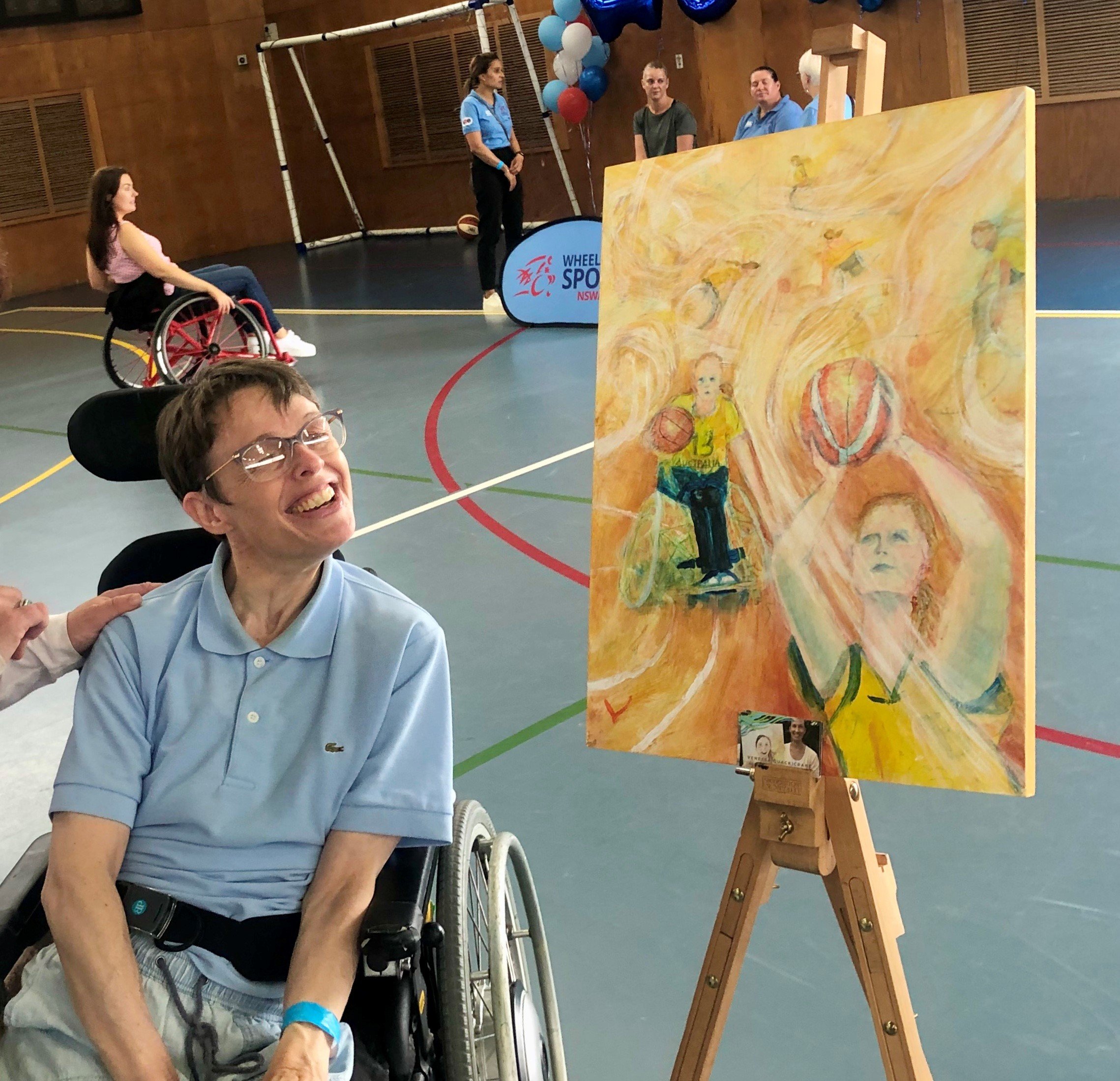 A young person in a light blue polo top and glasses sits in a wheelchair smiling on an indoor basketball court. Next to them is a yellow painting of people playing wheelchair basketball. Behind them is a person in a red wheelchair and spots officials standing to the side of the court with red, blue and white balloons