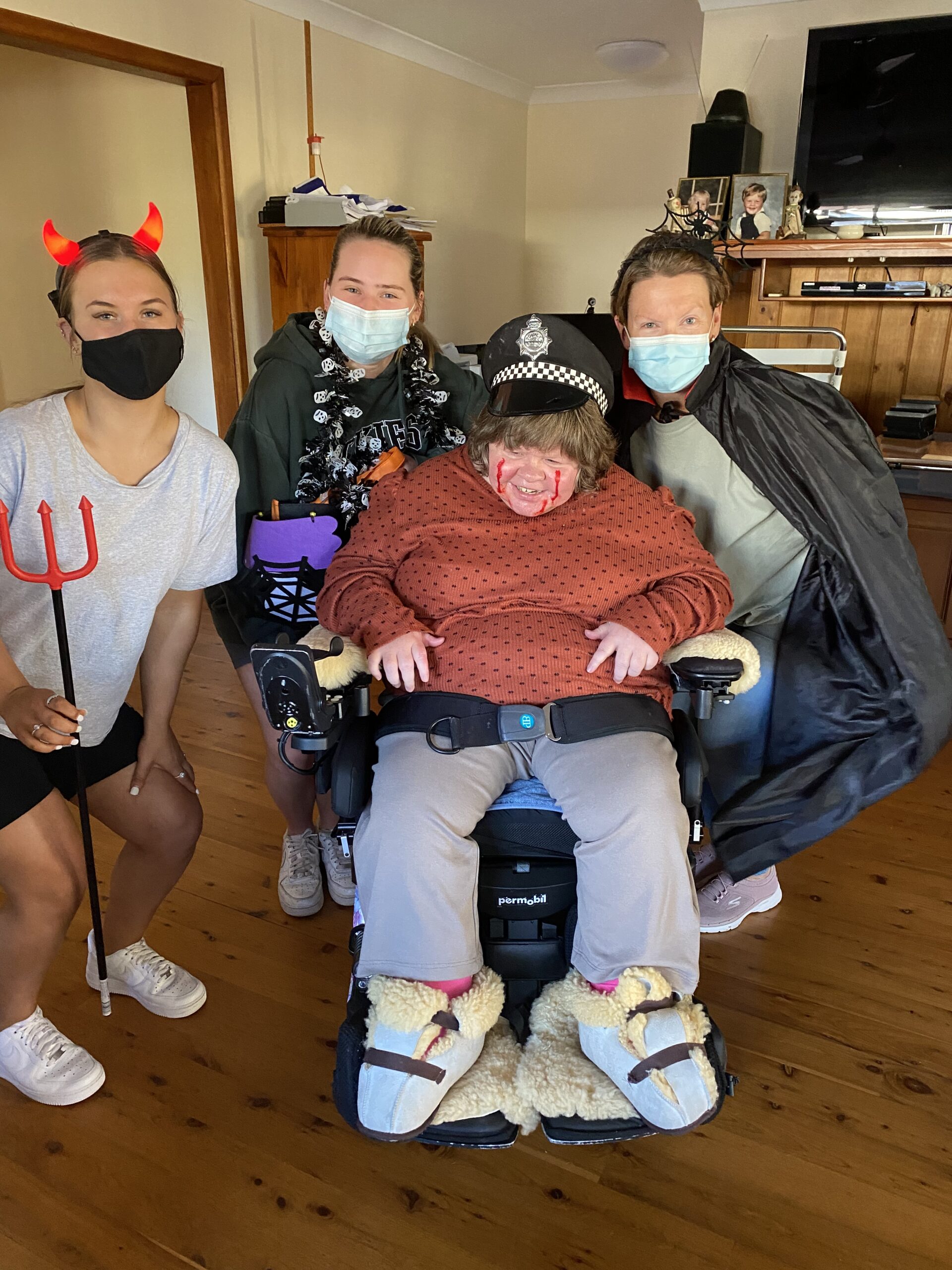 a person in a police hat, burnt red jumper and red blood-like face paint stains on their face sits in a power wheelchair with three people crouching behind them: one has a black face marks with red devils horns and red pitch fork, another has a blue surgicall mask and a cobwebbed-laden cape, the their has a blue surgical mask and a black cape and a cobweb poking out of their hair