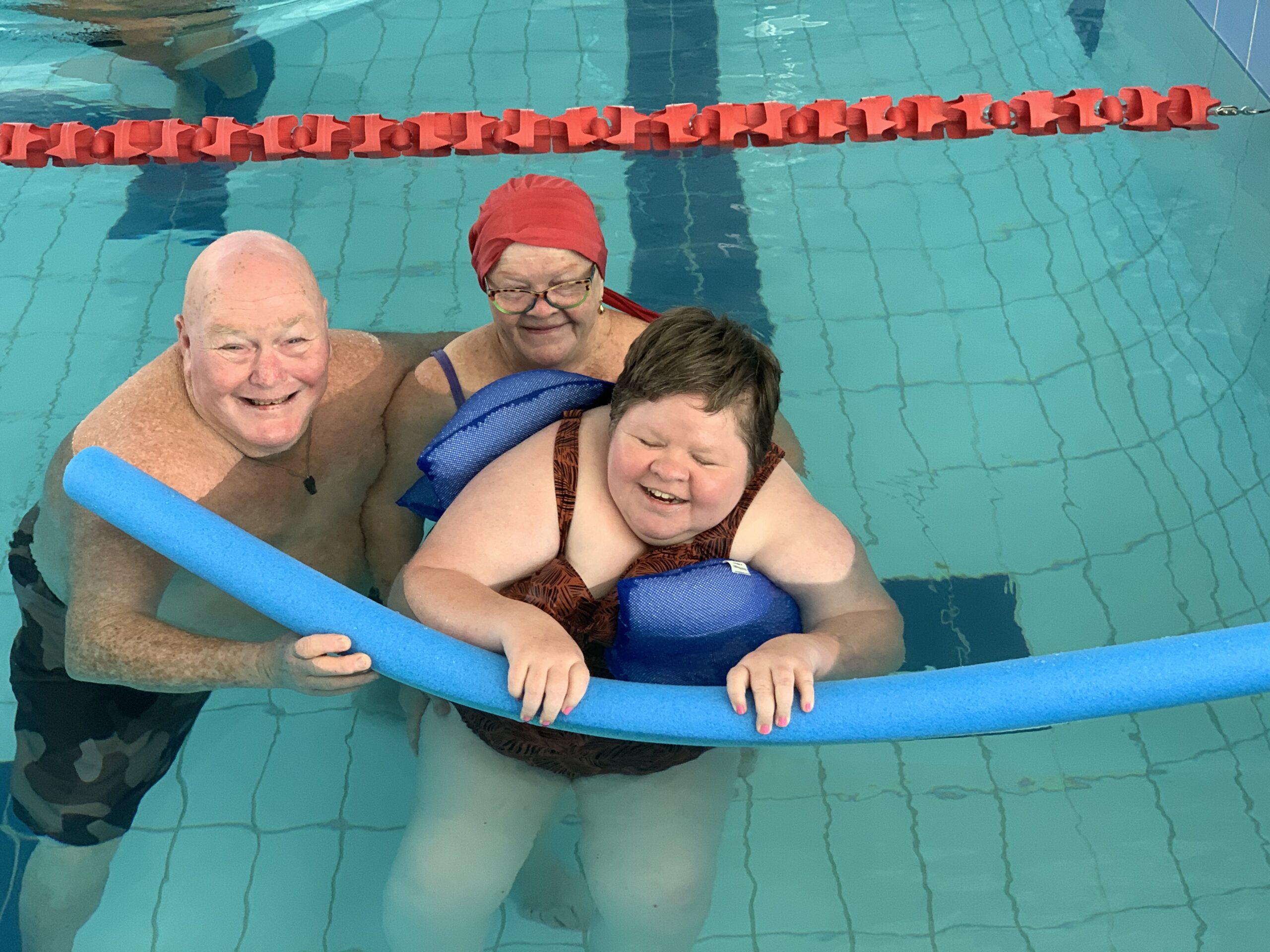 Three people smiling looking up as they stand in a pool: One person is wrapped in floatation beans and holds onto a noodle, a woman in a red cap stands behind holding onto the person in floatation beans and a man stands beside them both holding onto the noodle and his other arm wrapped around the woman in a red cap.