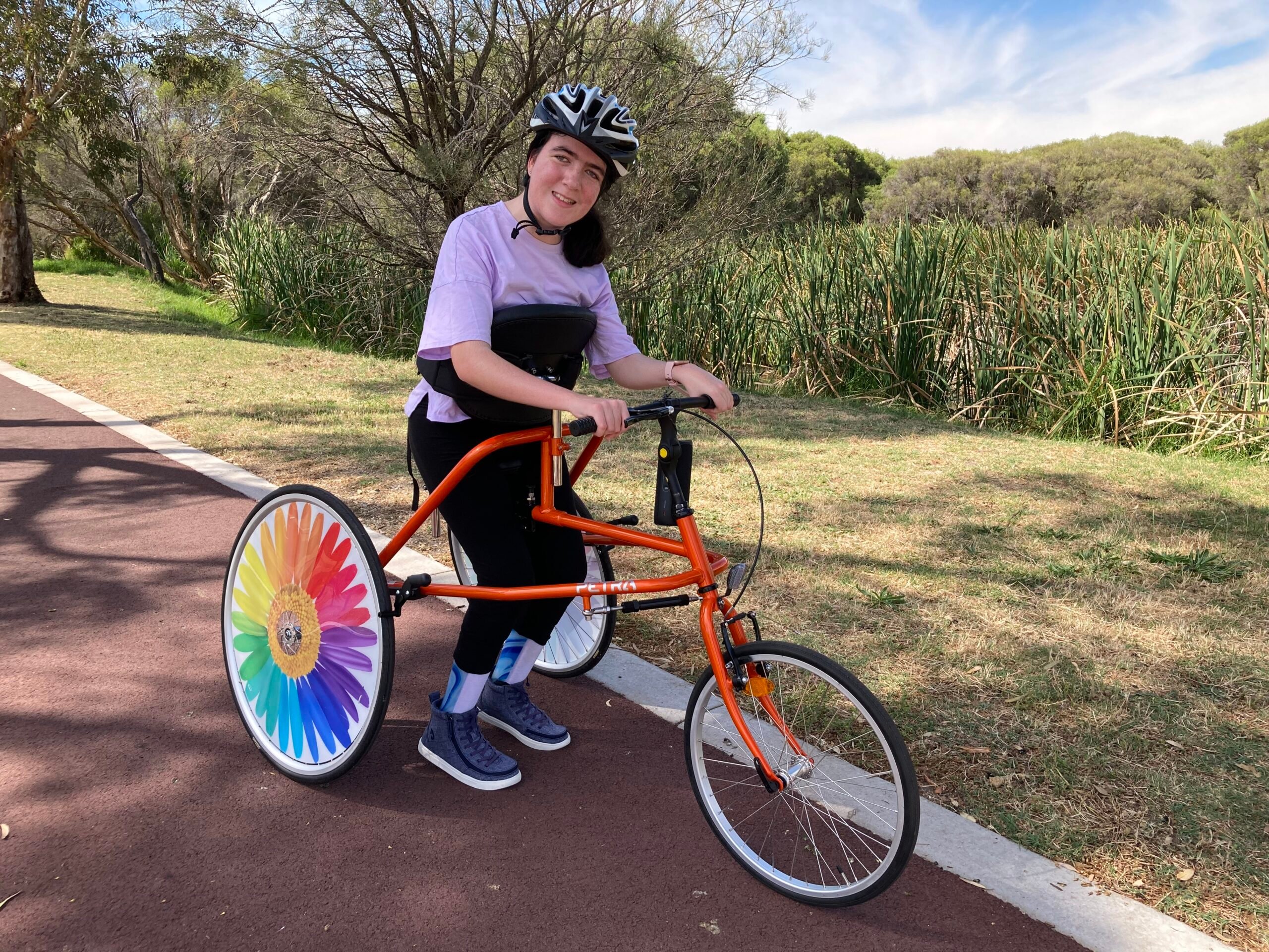 A young lady stands within a three-wheeled race walker with a bright rainbow rear wheel and helmet on her head. She is on a brown track with flat grass beside her and tall reeds and trees beyond in the background