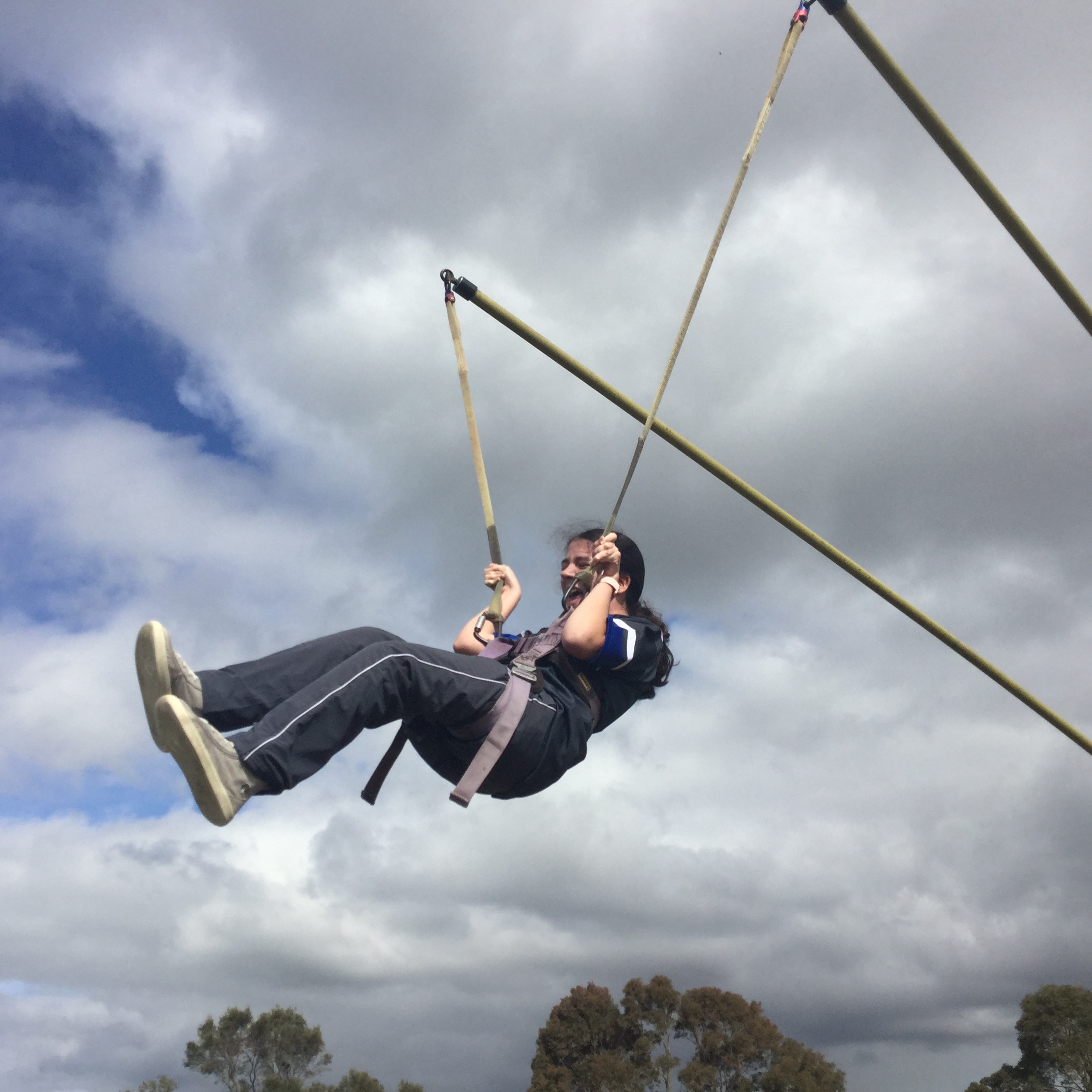 a young person sits in a harness smiling and holding on tightly to the cords coming from their harness. Two poles suspend the person off the ground and a white cloudy and blue sky are seen beyond them