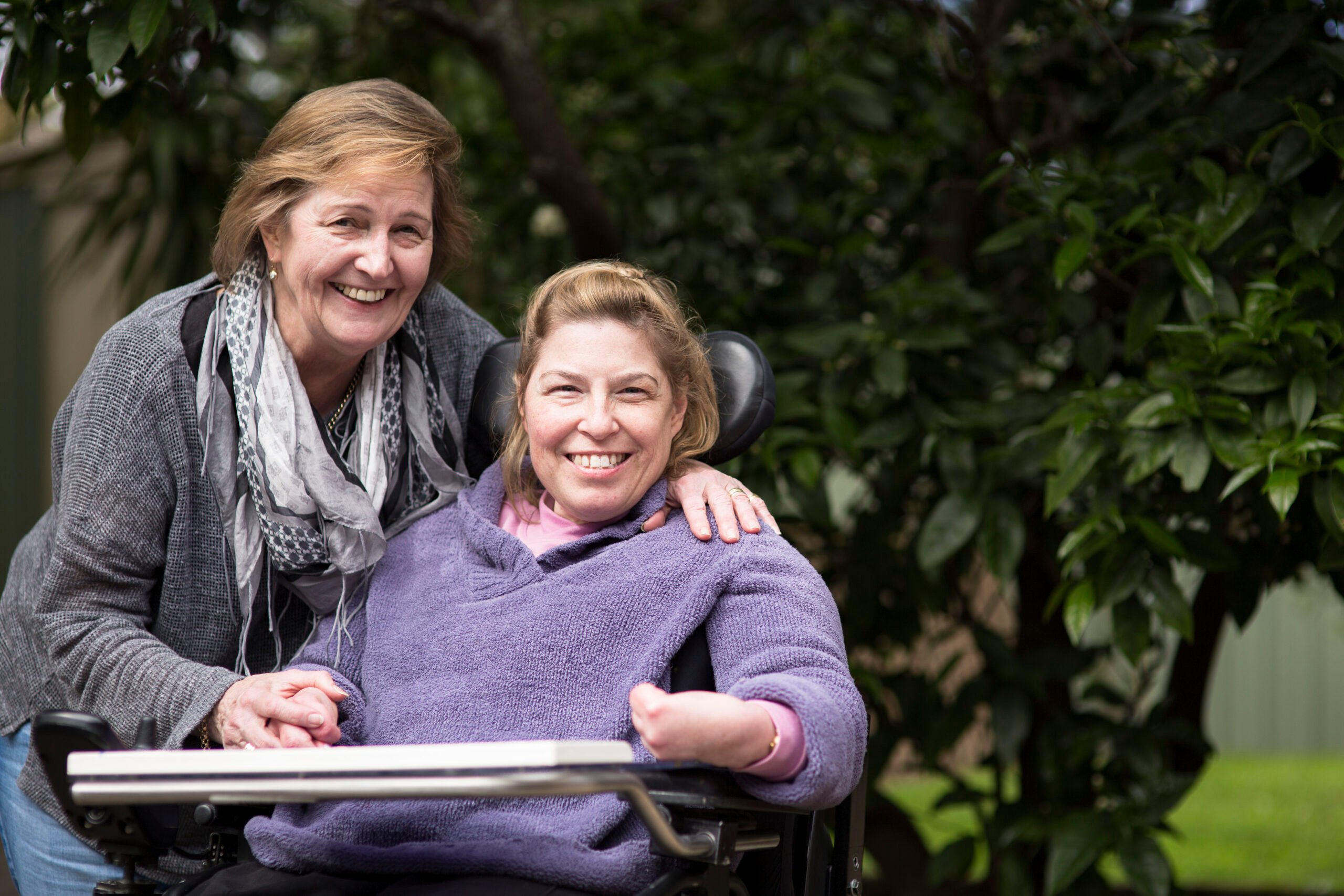 This image shows two women   smiling, located in a sunny area underneath some trees. One woman extends her arm to rest it on the shoulder of the other woman.
