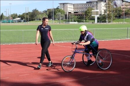 A young woman is using a running frame at an athletics track. Her running coach is walking alongside her.